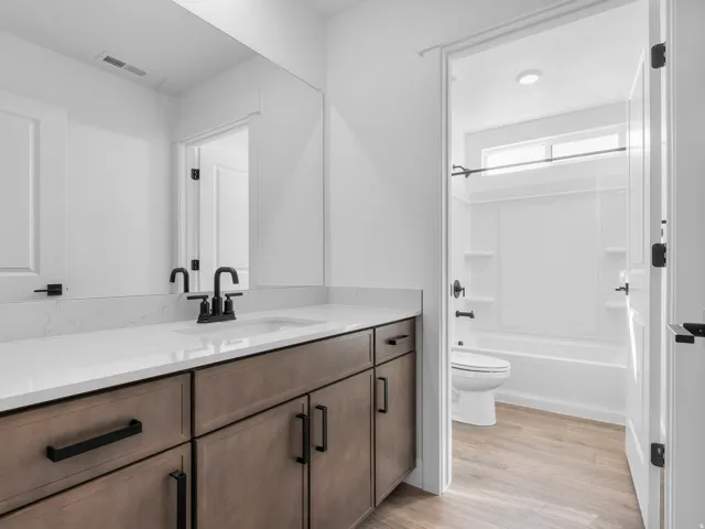 White countertop with integrated sink, black hardware, wood-finish cabinetry, full-width mirror, and a full bath with a window