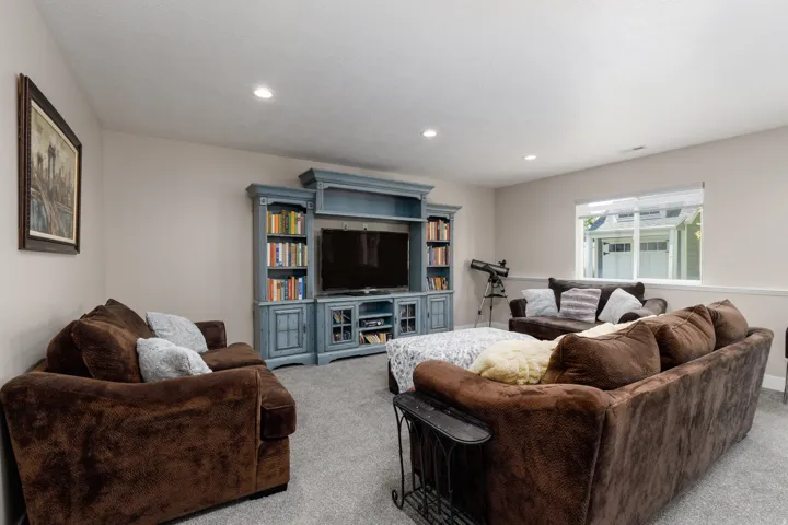 Spacious recreation room featuring dual brown upholstered seating areas, recessed ceiling lighting, carpet flooring, and a custom blue built-in entertainment center with shelving and media storage