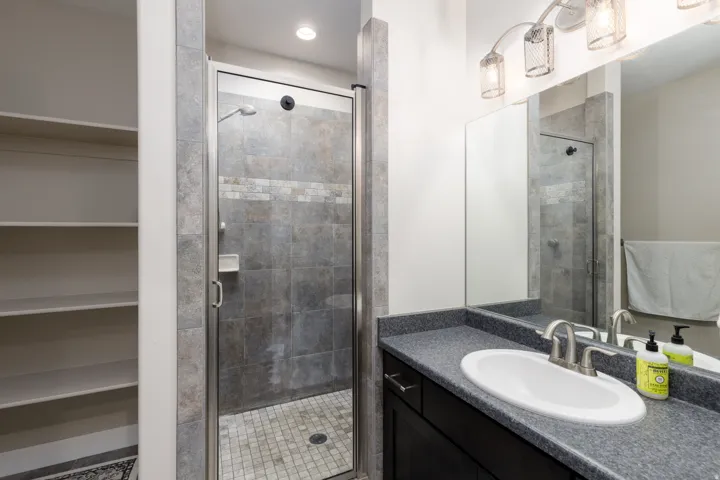 Walk-in shower with glass enclosure and tiled surround, vanity with integrated sink, dark cabinetry with brushed nickel hardware, and built-in shelving
