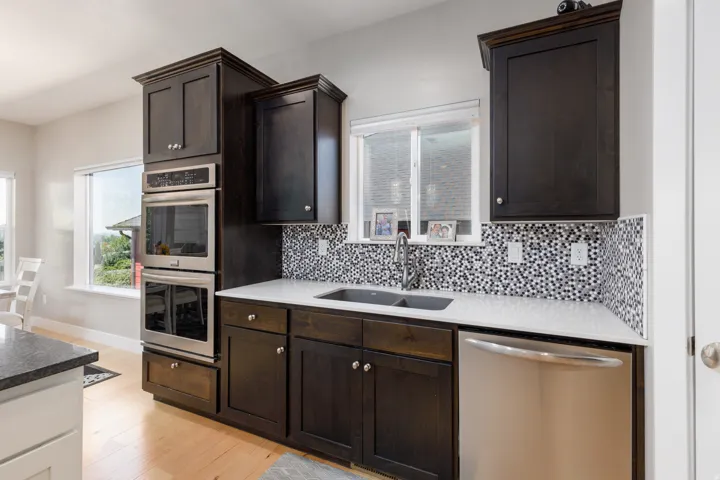 Dark wood-finish cabinetry, stainless steel double wall ovens, black and white mosaic tile backsplash, solid surface countertops, and light wood-finish flooring
