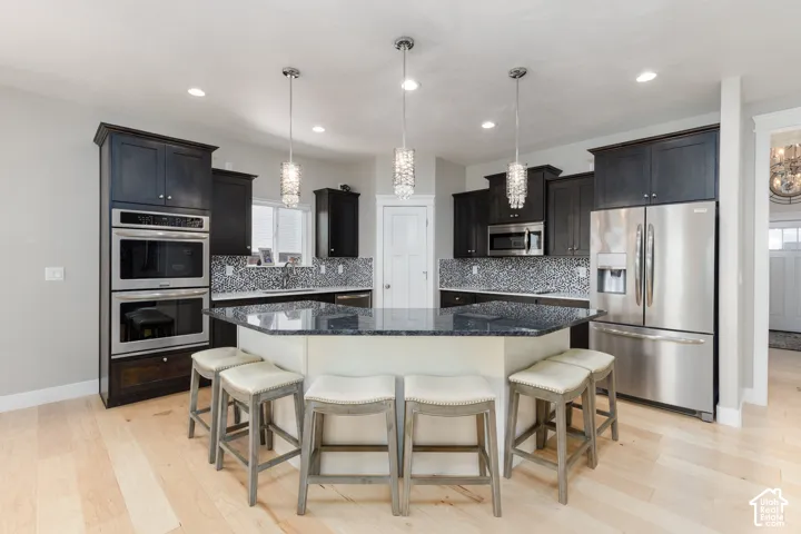 Kitchen featuring dark cabinetry, stainless steel appliances, a tile backsplash, light wood-finish flooring, and a multi-sided island with pendant lighting
