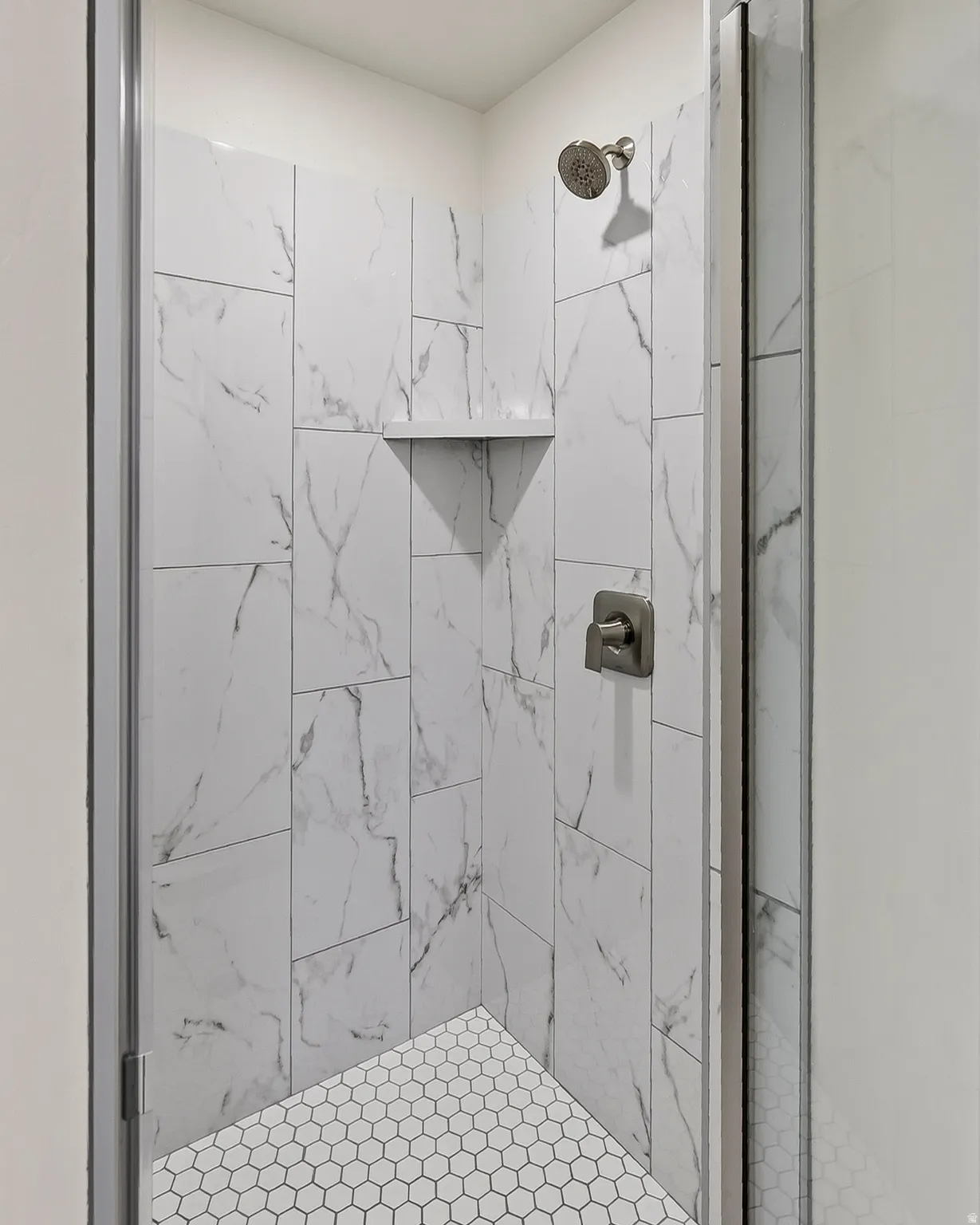 Enclosed shower featuring white marble-look wall tiles, hexagonal floor tiles, a built-in shower shelf, and brushed nickel fixtures
