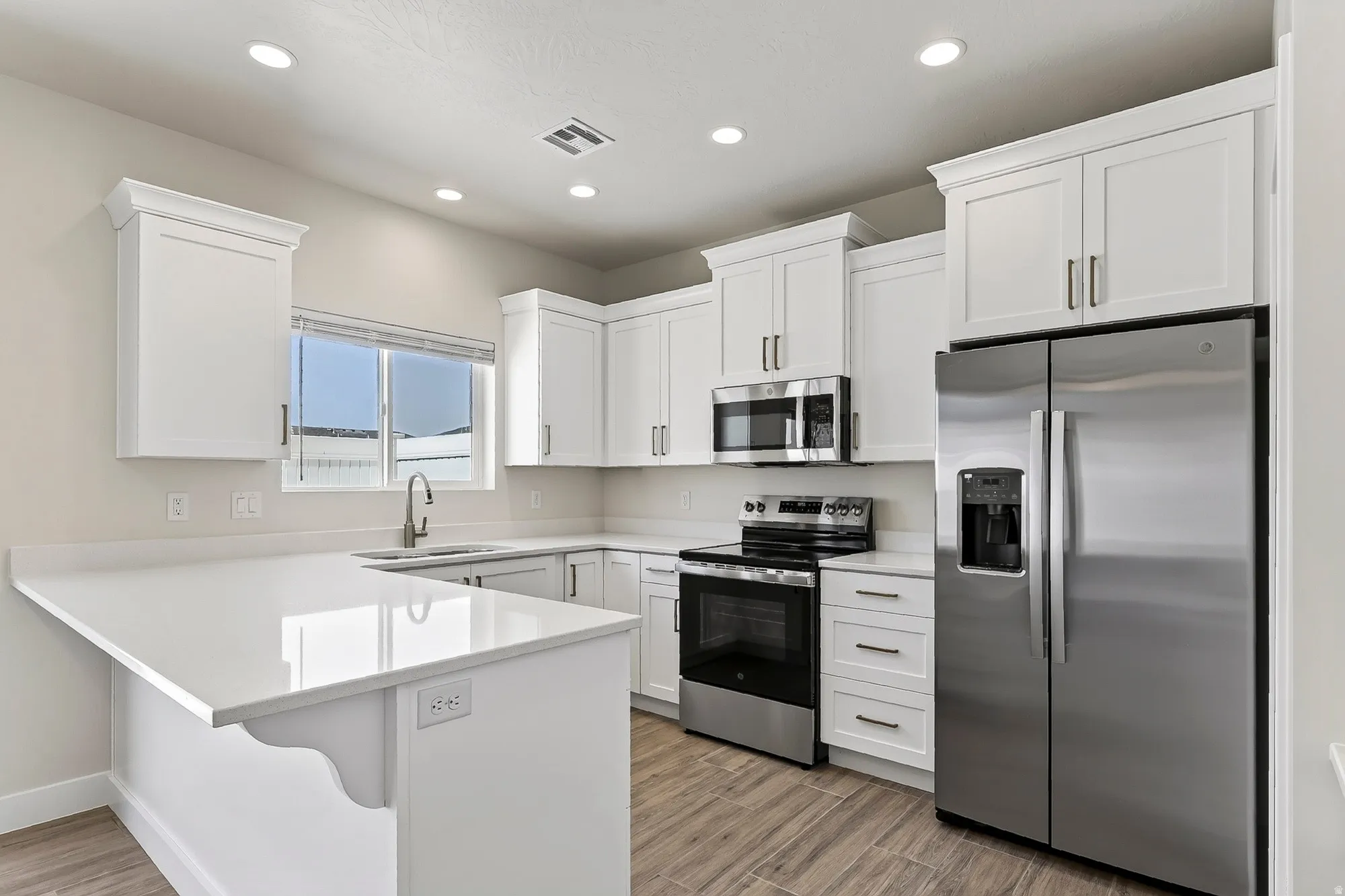 White shaker cabinetry, stainless steel appliances, light-colored countertops, wood-finish flooring, and recessed lighting