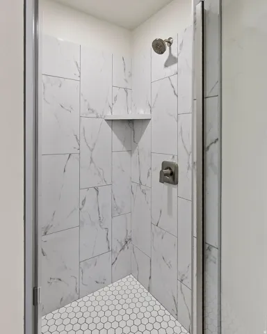 Enclosed shower featuring white marble-look wall tiles, hexagonal floor tiles, a built-in shower shelf, and brushed nickel fixtures