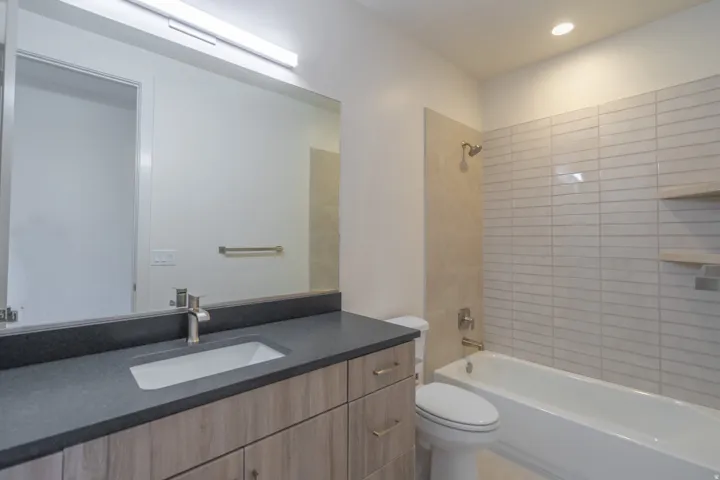 Bathroom featuring a wood-finish vanity with a dark countertop and integrated sink, a large mirror, and a white bathtub with light-toned tiled surround