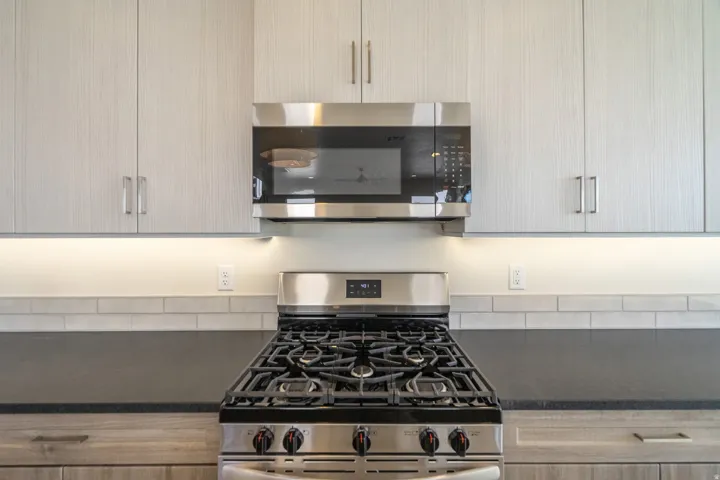 Kitchen featuring a stainless steel gas range with grates, coordinated microwave, light wood-grain cabinetry, dark countertops, and a subway tile backsplash