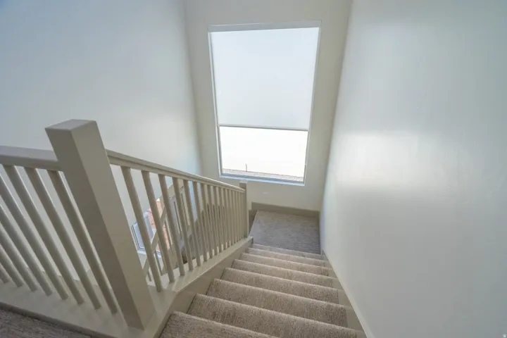 Carpeted staircase with light-toned railings and a large, tall window featuring a roller shade