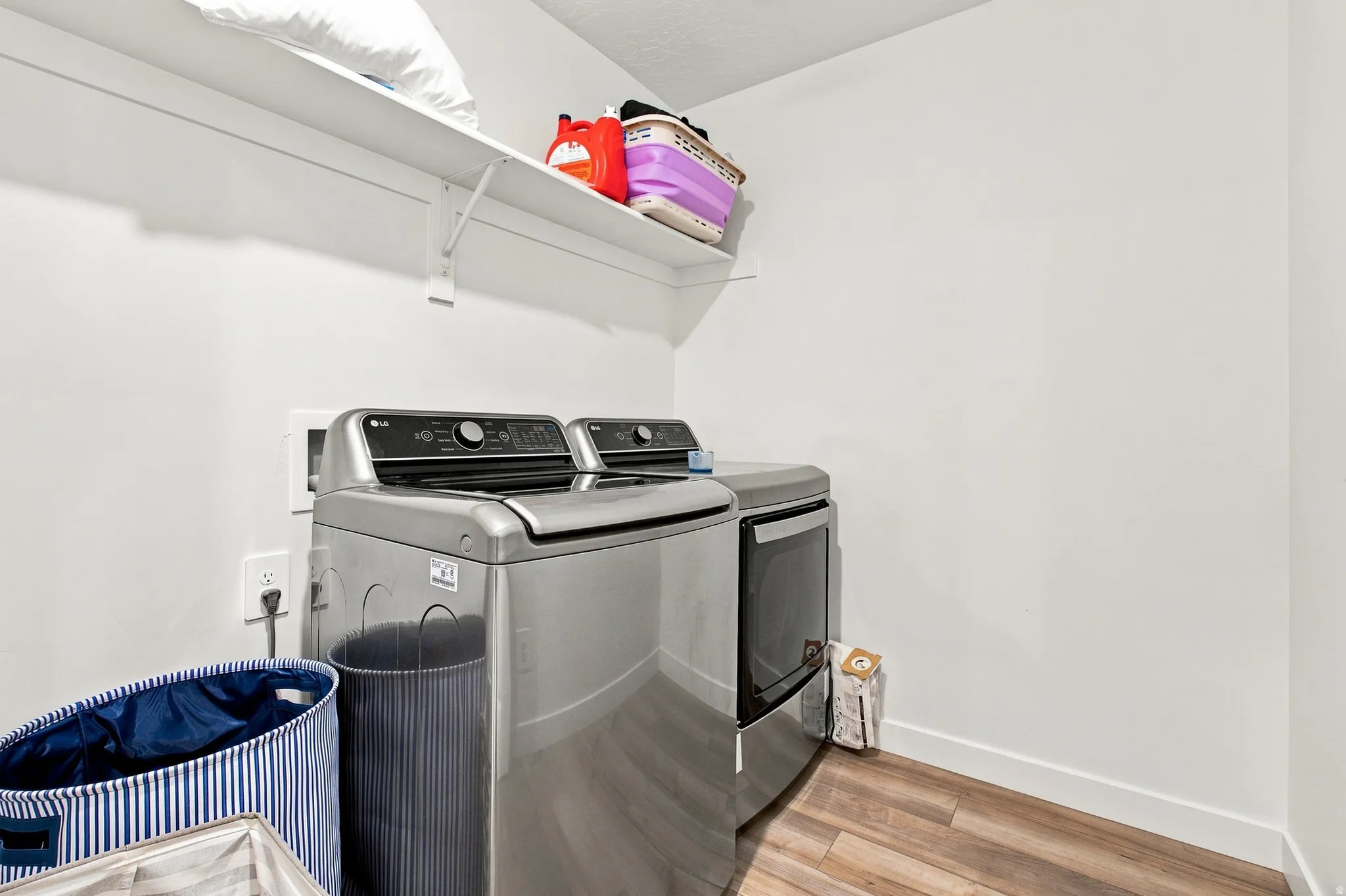 Laundry area featuring durable wood-finish flooring, a wall-mounted shelving unit, and white walls