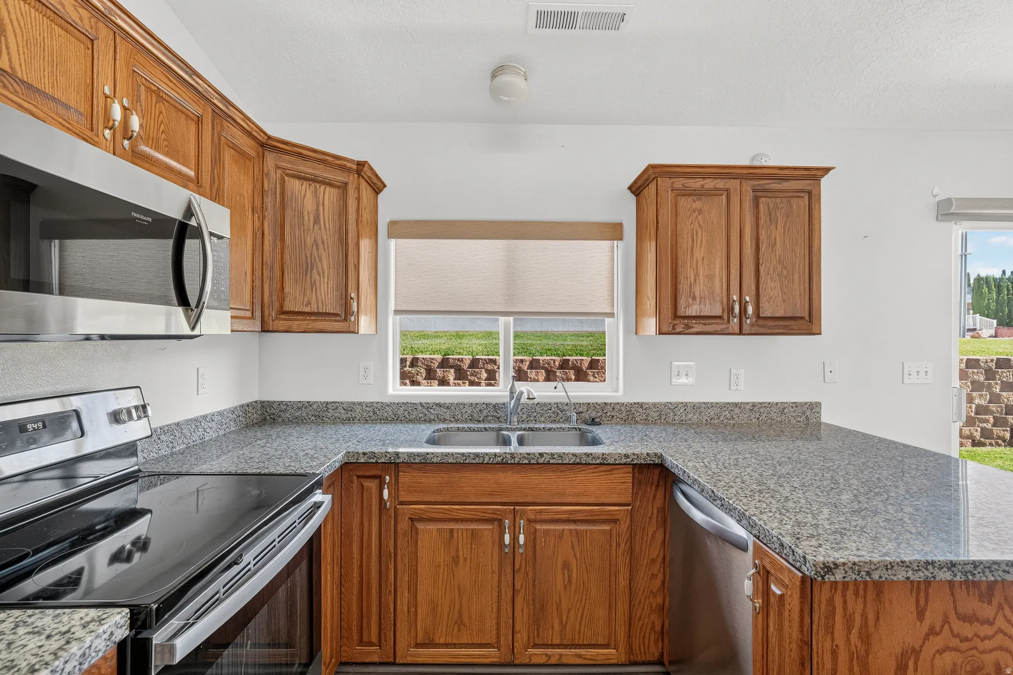 Warm wood cabinetry and speckled granite countertops define this well-appointed kitchen, featuring stainless steel appliances, a dual-basin sink with chrome fixtures, and ample counter workspace