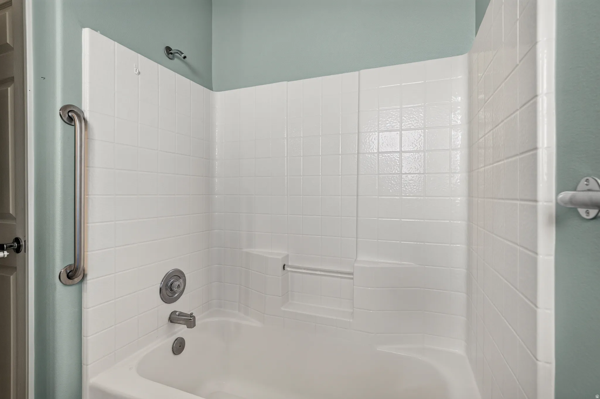 White tiled tub and shower combination featuring a brushed nickel grab bar, integrated shelving, and a coordinating shower head fixture
