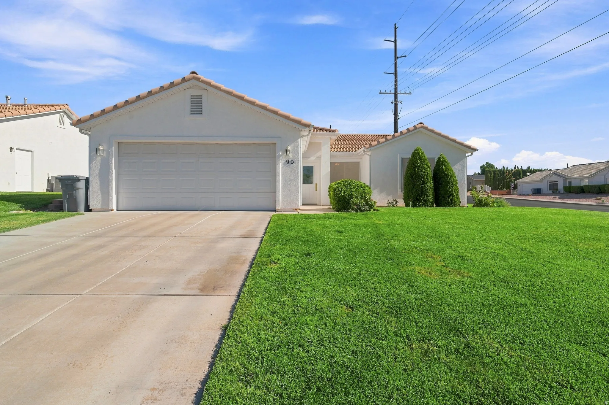 Contemporary stucco exterior with a terracotta tile roof, attached two-car garage, concrete driveway, and a spacious front lawn with mature shrubbery