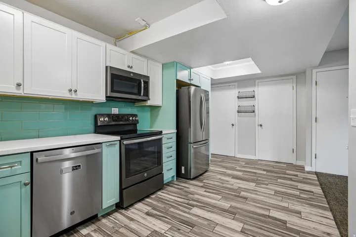 Stainless steel appliances, two-tone cabinetry with white upper and teal lower units, subway tile backsplash, white countertops, and wood-finish flooring