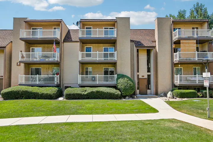 Modern multi-unit residential complex featuring clean architectural lines, stacked balconies with white railings across three stories, warm-toned exterior façade, manicured landscaping with mature shrubs and green lawn frontage, and multiple ground-level