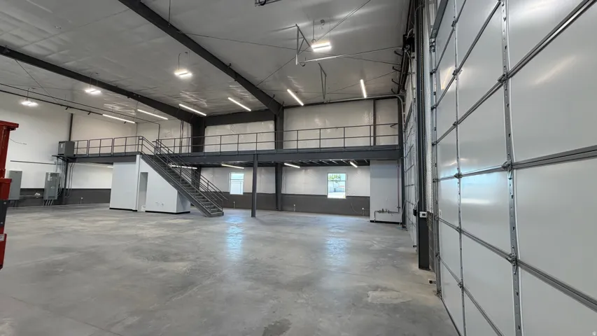 Open-concept commercial space featuring high ceilings, exposed structural beams, a second-story mezzanine with metal railing, and a large roll-up door