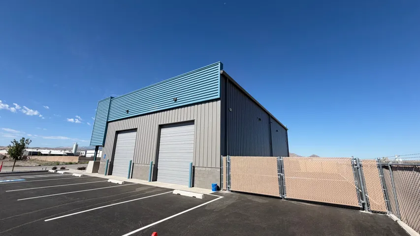 Metal-clad building with two roll-up doors, a paved parking lot, and a chain-link fence with privacy slats