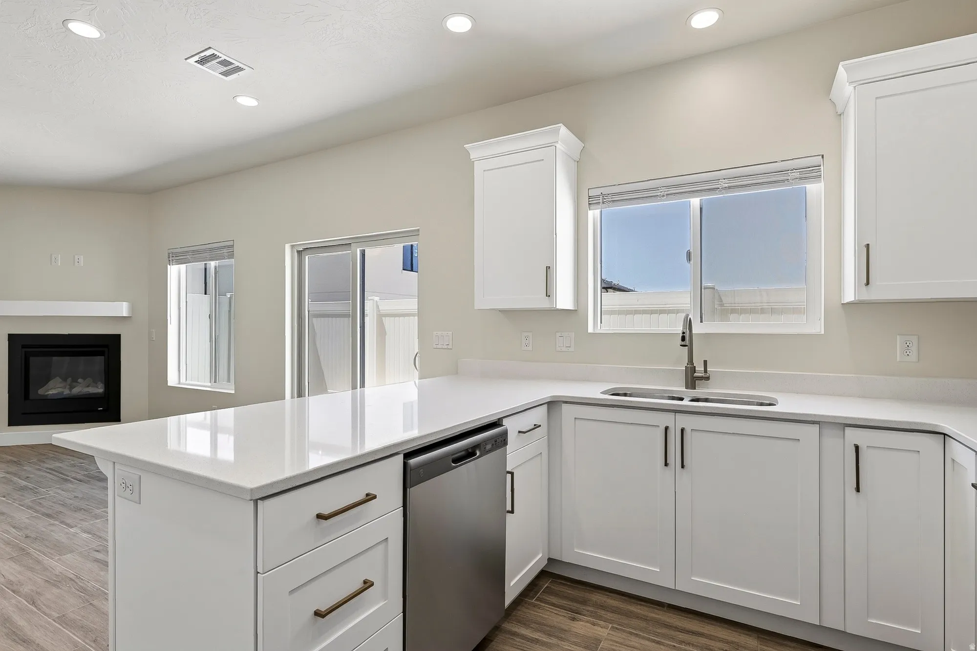 White shaker-style cabinetry with bronze hardware, stainless steel dishwasher, undermount sink with gooseneck faucet, light-toned countertops, and wood-finish flooring