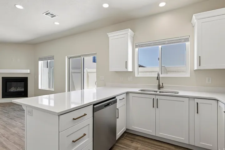 White shaker-style cabinetry with bronze hardware, stainless steel dishwasher, undermount sink with gooseneck faucet, light-toned countertops, and wood-finish flooring