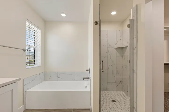 Spa-inspired primary bathroom featuring a soaking tub with tile surround, a frameless glass-enclosed walk-in shower with mosaic tile flooring and built-in niche, dual vanity with quartz countertop, recessed lighting, and a transom window providing natural