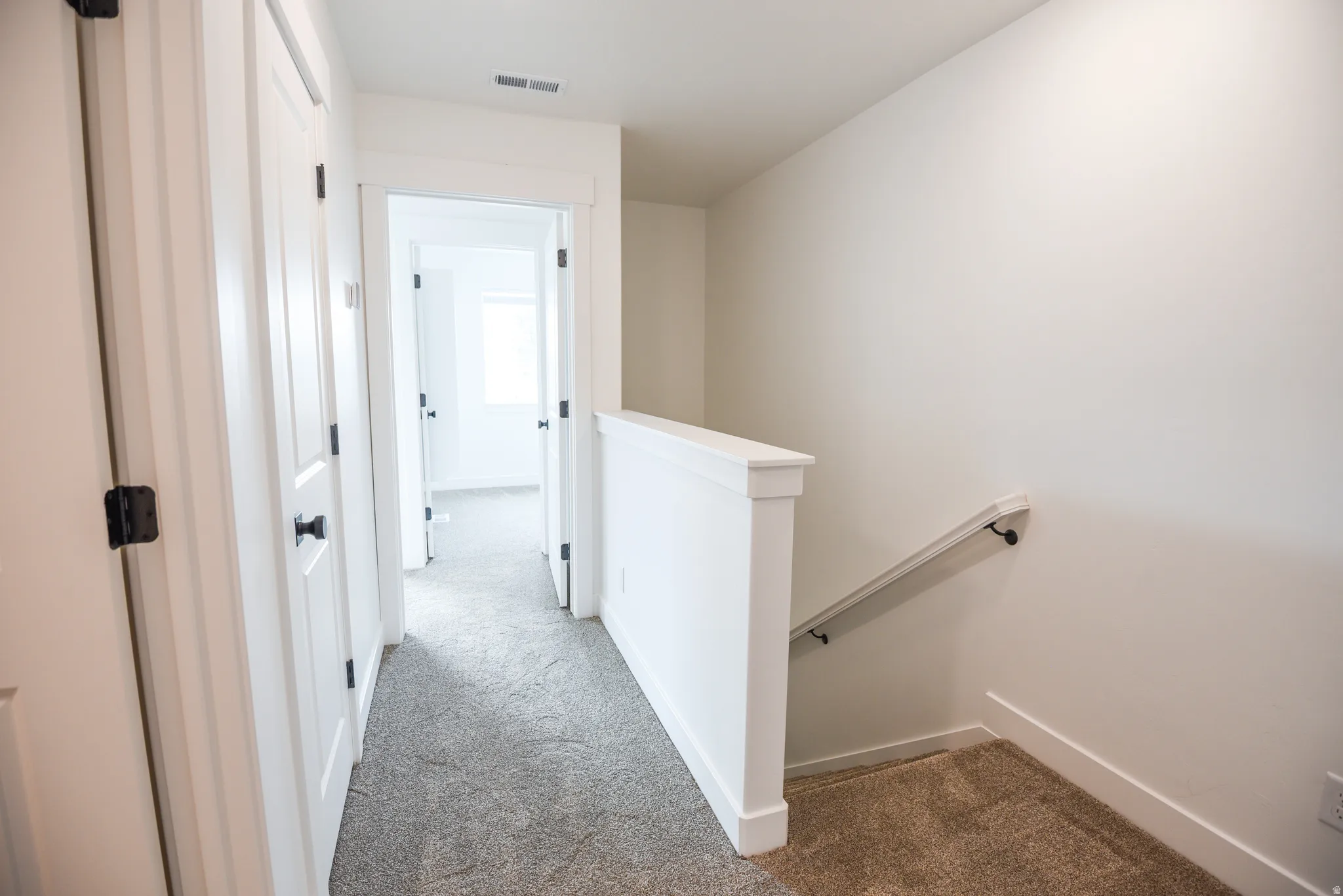 Hallway with gray carpeting, white walls, white doors with matte black hardware, and a white stair railing with a black handrail