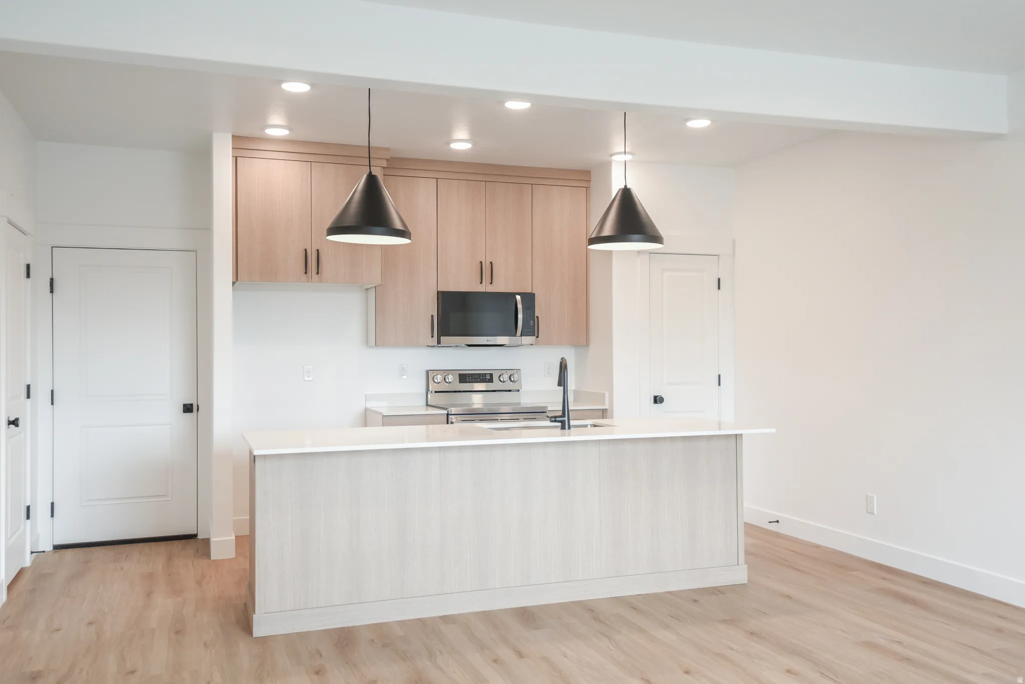Kitchen island with an undermount sink and matte black faucet, light wood-finish cabinetry, stainless steel appliances, recessed lighting, and wood-finish flooring