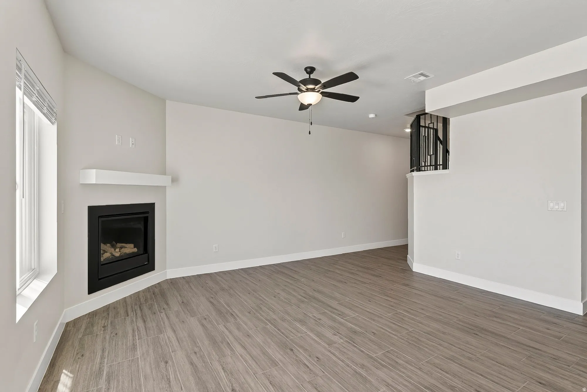 Angled fireplace with a white mantel, wood-finish flooring, ceiling fan with integrated lighting, and a partial black metal stair railing