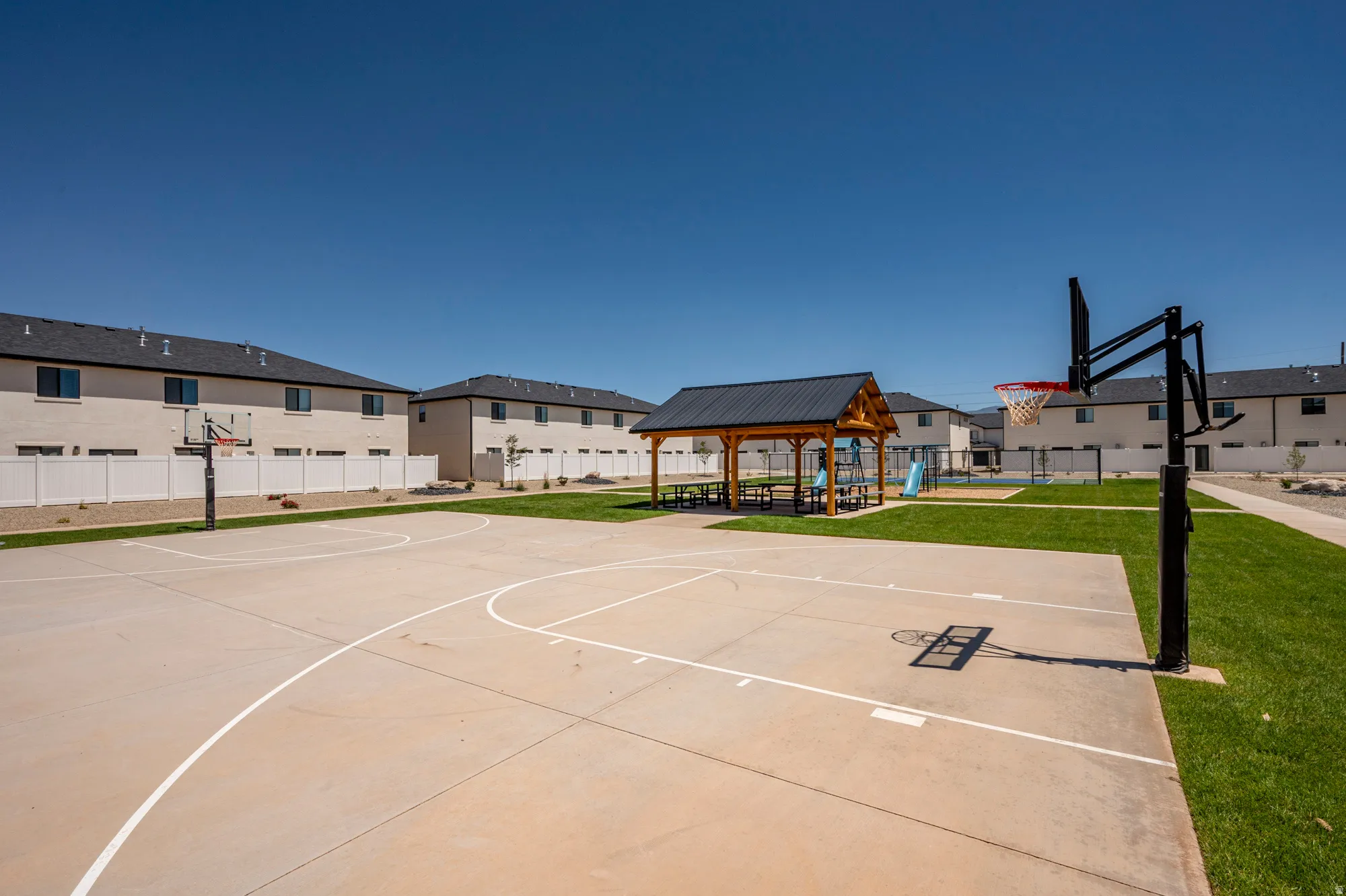 Outdoor recreational space featuring a concrete basketball court, a covered picnic area with a dark metal roof, and manicured green lawns