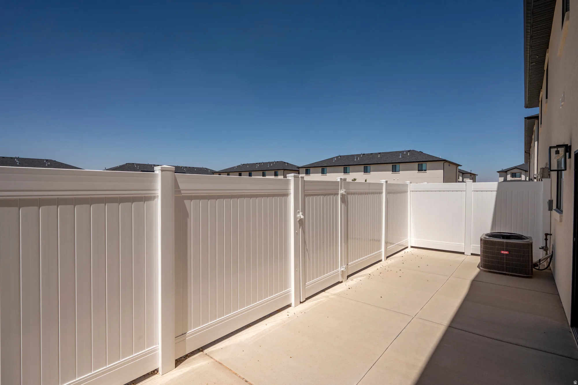 Paved concrete patio area featuring white fencing, an exterior wall light fixture, and an HVAC unit