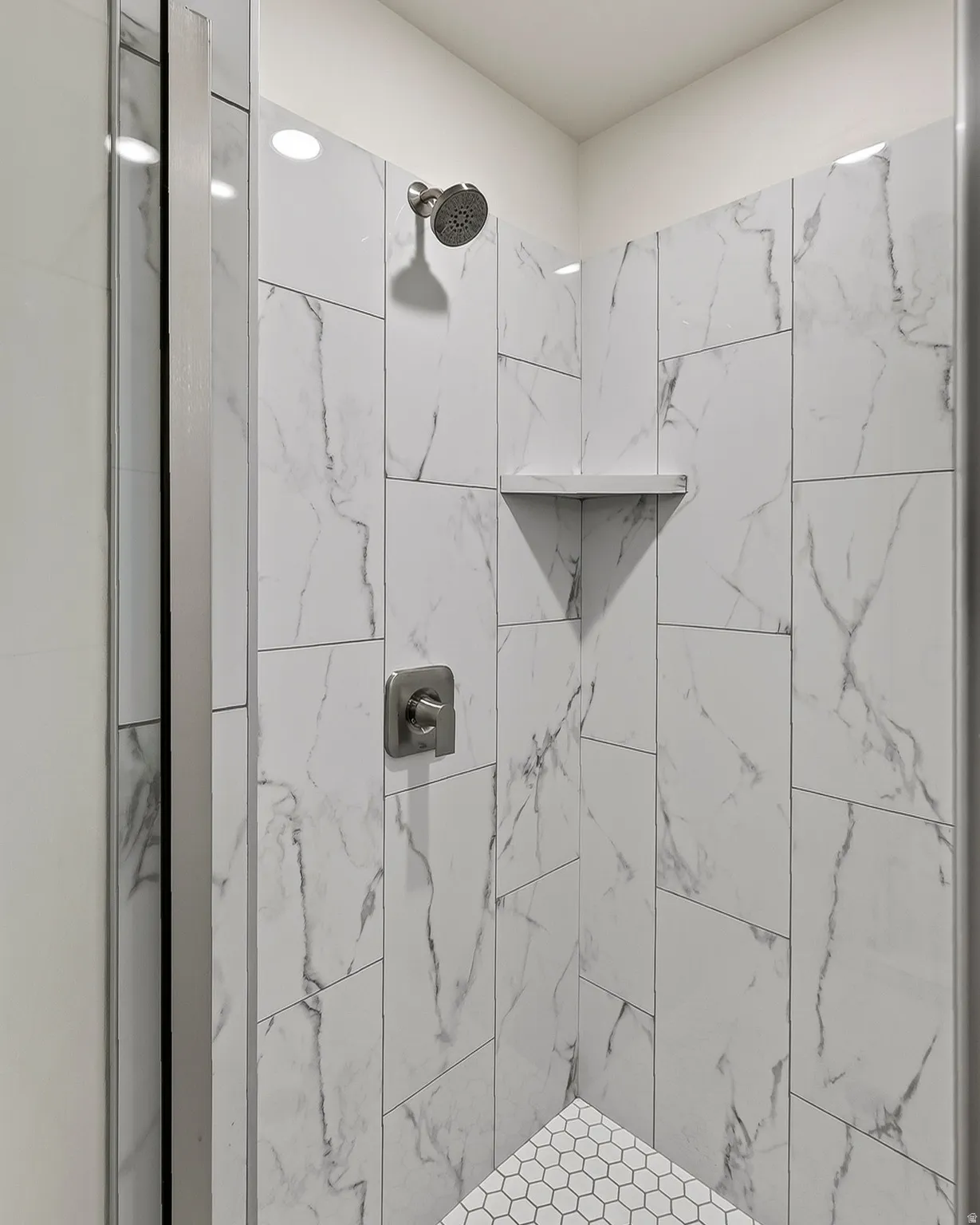 Shower enclosure featuring white marble-look wall tiling, a built-in corner shelf, and hexagon mosaic floor tiling