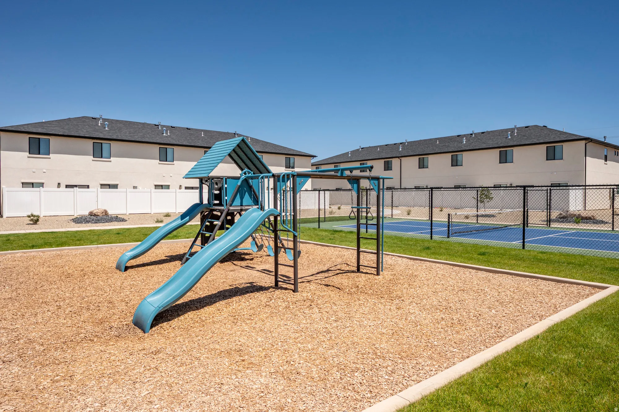 Community playground with wood chip surfacing, turf grass areas, and a multi-sport court