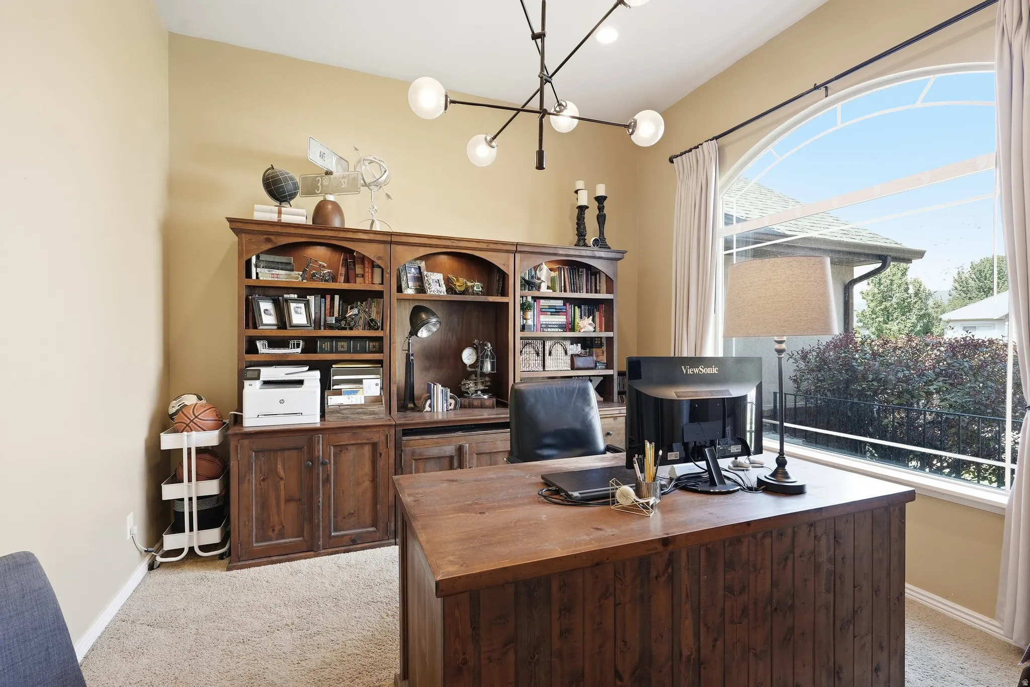Built-in wood-finish cabinetry, arched window with white trim, modern chandelier, wood-finish desk, and carpeting