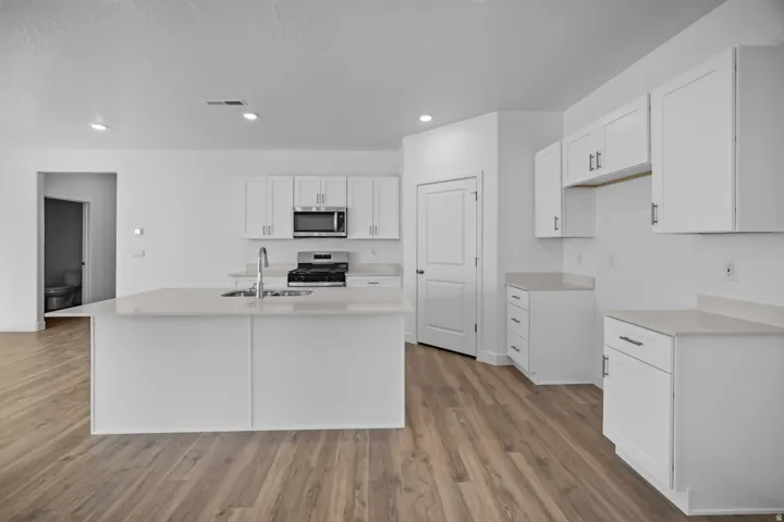 Central island with an undermount sink, white cabinetry, integrated stainless steel appliances, light-toned countertops, and wood-finish flooring