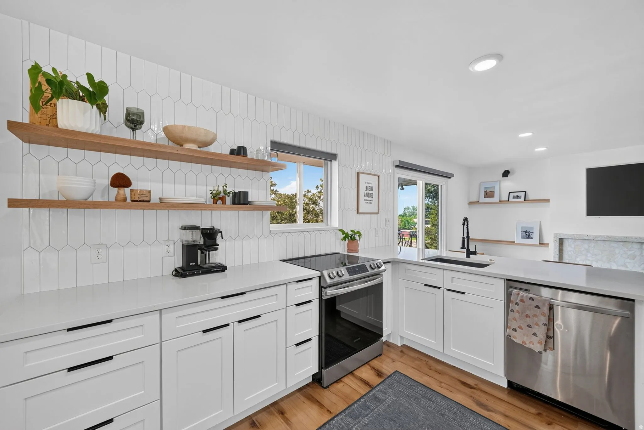 Modern kitchen featuring white cabinetry with integrated hardware, stainless steel appliances including a gas range and dishwasher, quartz countertops, a undermount sink with contemporary faucet, white subway tile backsplash, floating wood shelving, and w