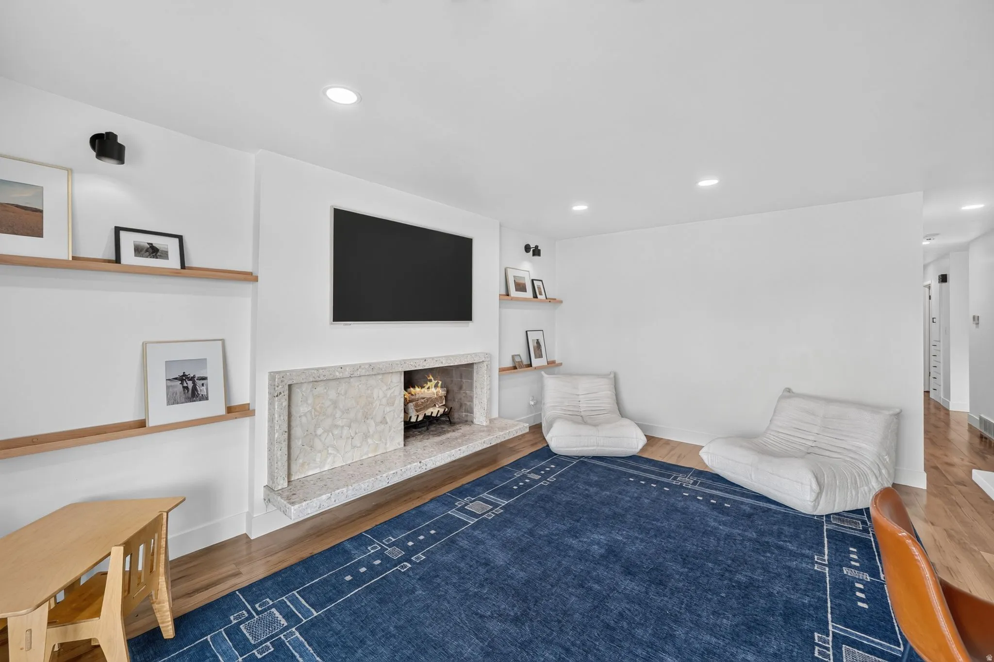 Modern recreation room featuring a wall-mounted flat-screen television, linear fireplace with stone surround and wood mantel, hardwood flooring, recessed ceiling lighting, built-in shelving with wood-finish frames, and a geometric patterned area rug in na