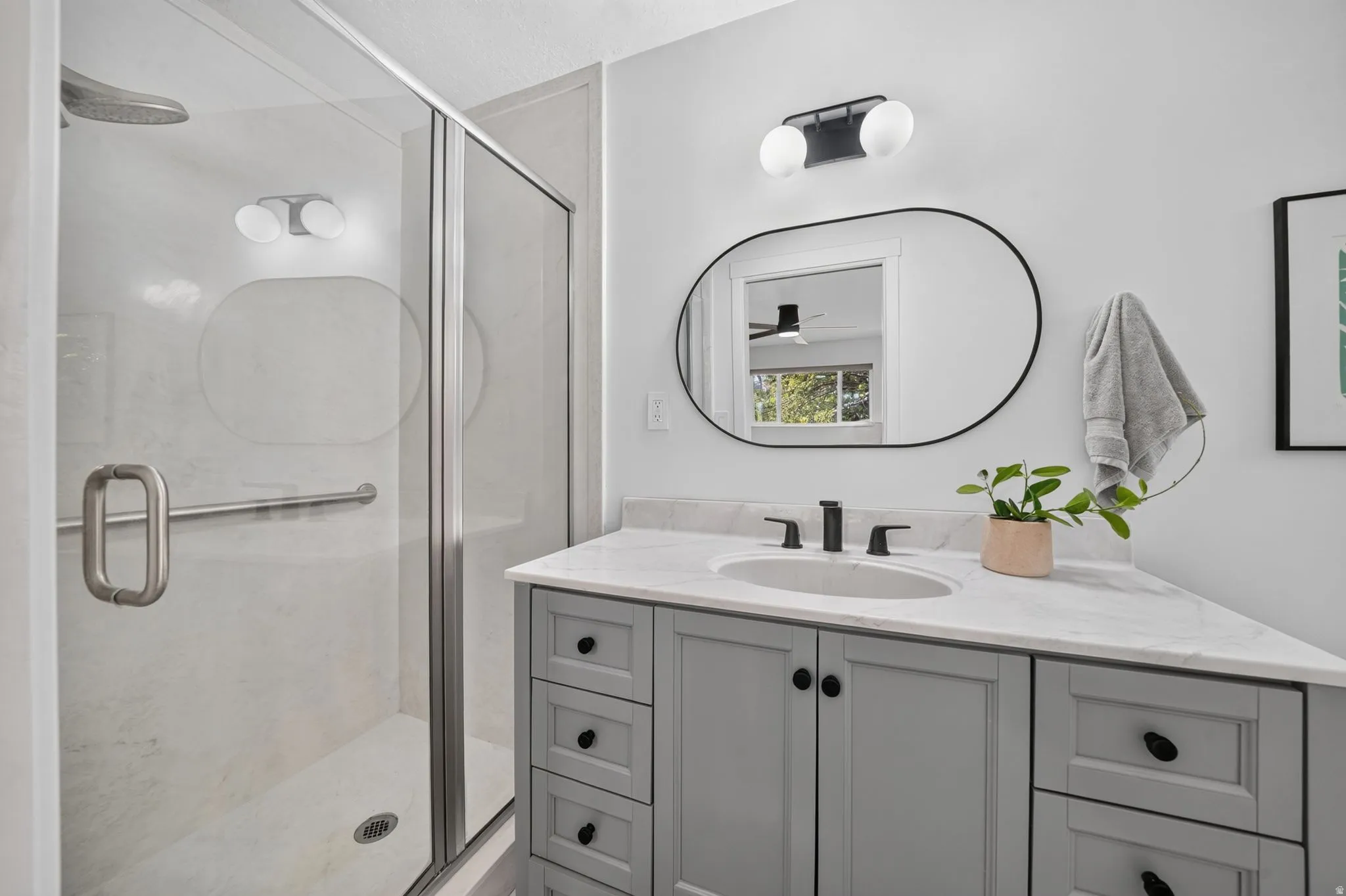 Frameless glass shower enclosure with a rain shower head, light grey vanity with a white countertop, oval vanity mirror, and modern wall-mounted light fixture