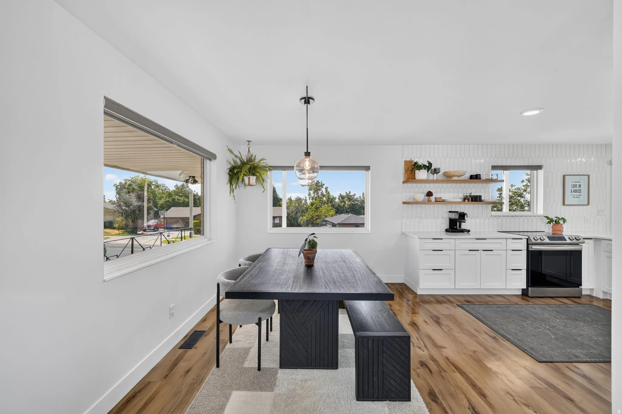 Open-concept kitchen and dining area featuring white cabinetry with integrated appliances, wood-finish flooring throughout, a substantial dining table with bench seating, pendant lighting, and expansive windows framing natural light and outdoor scenery