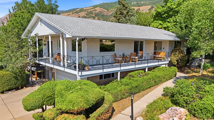 Exterior featuring white brick siding, a large elevated deck with decorative metal railings, a paved driveway, and mature landscaping