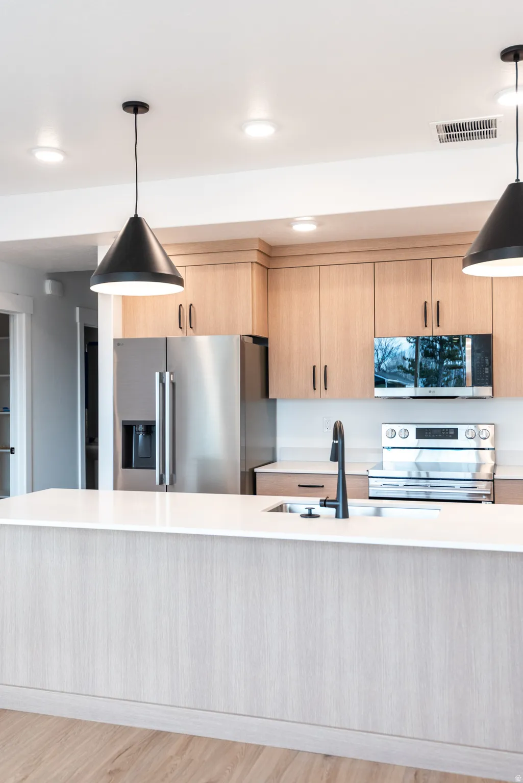 Kitchen featuring light wood-finish cabinetry, stainless steel appliances, a large island with white countertops, a black gooseneck faucet, and recessed lighting