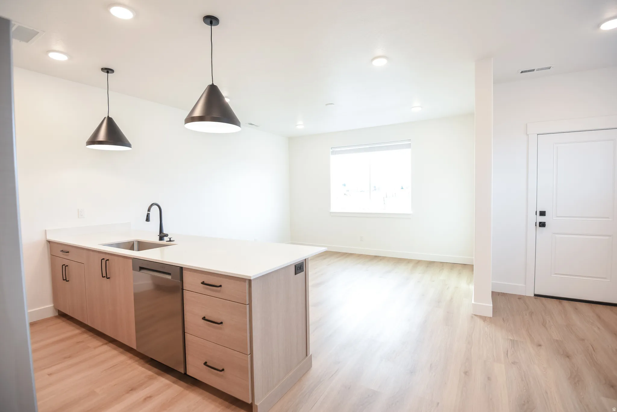 Kitchen island with a white countertop and integrated sink, stainless steel dishwasher, light wood-finish cabinetry, and black pendant lighting