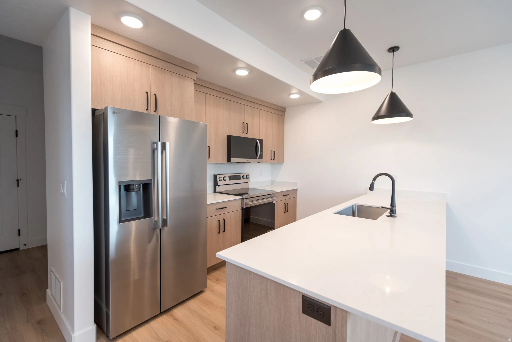 Contemporary kitchen featuring light wood-finish cabinetry, stainless steel appliances, white countertops, and a kitchen island with a built-in sink and matte black faucet
