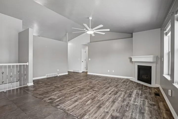 Spacious open-concept living area featuring wood-finish flooring, ceiling fan with integrated lighting, fireplace with surround, and abundant natural light from multiple windows
