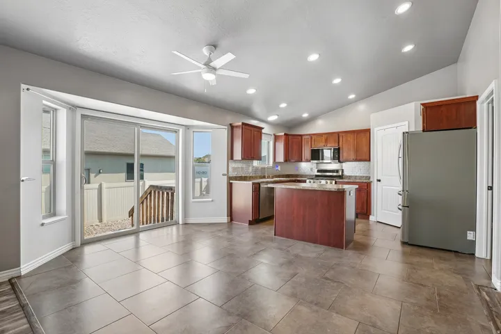 Spacious open-concept kitchen and dining area featuring rich wood cabinetry, stainless steel appliances, a large center island with seating, recessed and ceiling fan lighting, tile flooring, and sliding glass doors opening to an outdoor deck with scenic w