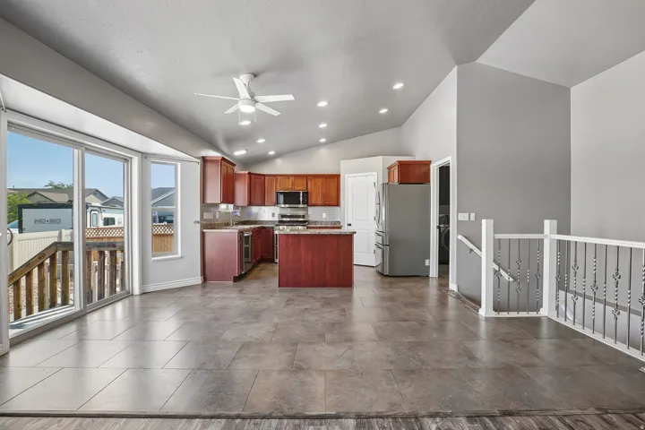 Open-concept kitchen and living area featuring stainless steel appliances, wood-finish cabinetry with island workspace, polished concrete flooring, ceiling fan with recessed lighting, sliding glass doors with deck access, and waterfront views through expa