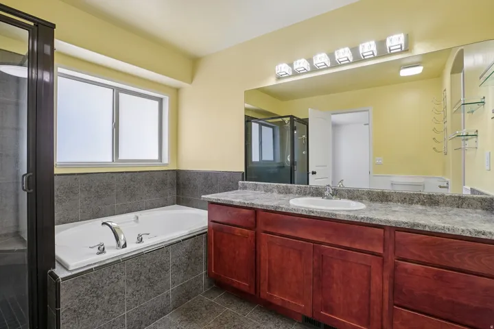 Built-in soaking tub, separate glass-enclosed shower, wood-finish vanity with an integrated sink, broad wall mirror, and tiled flooring