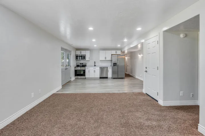 Open-concept living and kitchen area featuring neutral carpeting and wood-finish flooring, recessed ceiling lighting, white cabinetry with integrated stainless steel appliances, and ample counter space