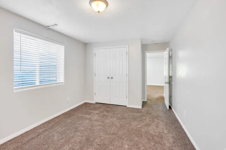 Spacious bedroom featuring neutral wall finishes, plush carpet flooring, and abundant natural light from a large window with horizontal blinds