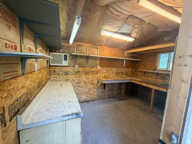 Workshop space featuring extensive built-in shelving, a large L-shaped workbench, a dedicated work surface, two fluorescent light fixtures, and a south facing window and air conditioner.