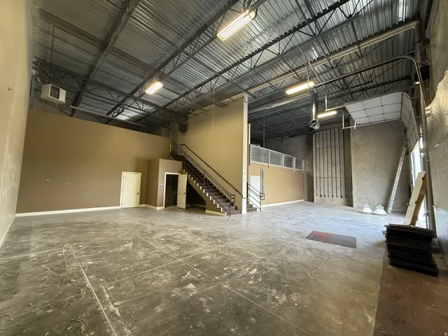 Expansive open-concept interior with concrete flooring and high ceilings, featuring a steel staircase with railing, multiple fluorescent lighting fixtures, and a large roll-up door