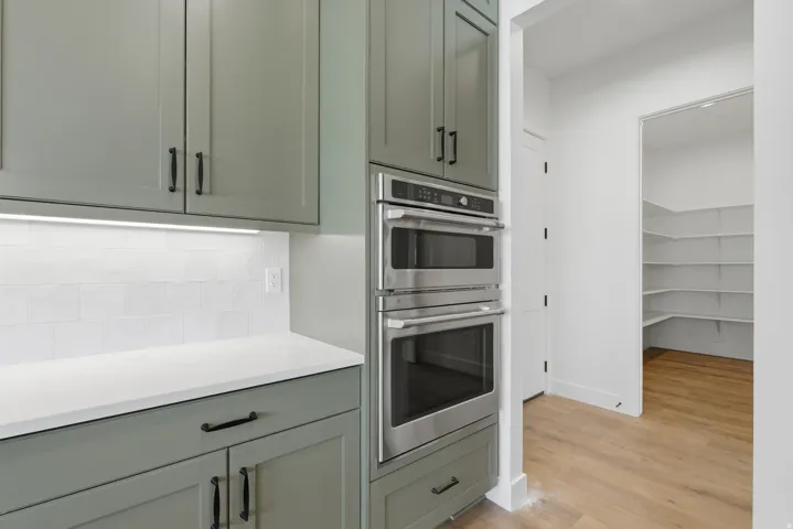 Sage green cabinetry with black hardware, white subway tile backsplash, under-cabinet lighting, and stainless steel double oven