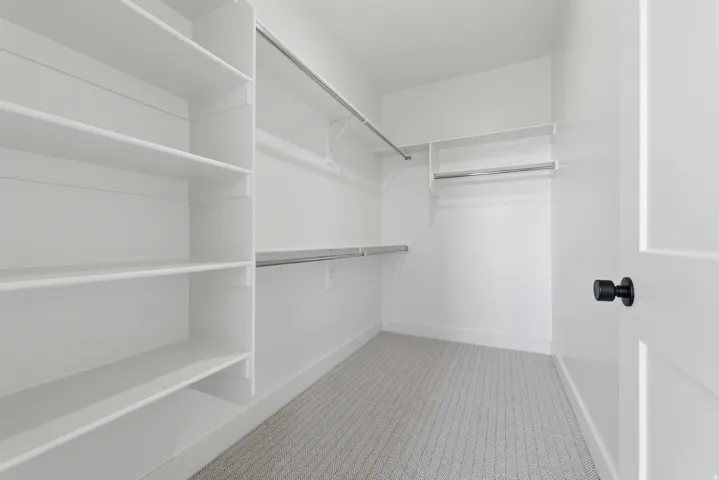 Walk-in closet featuring white built-in shelving, multiple hanging rods, and light-toned patterned carpeting