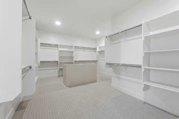 Primary suite. Spacious walk-in closet featuring extensive built-in shelving, multiple hanging rods, a central storage island, recessed lighting, and patterned carpeting
